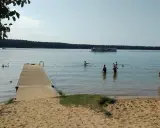 Słoneczna augustów plaża z pomostem, łabędziami i ludźmi kąpiącymi się w jeziorze. Na wodzie pływa statek wycieczkowy.