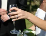 Dłoń panny młodej z pierścionkiem, wznosząca kieliszek szampana. W tle pan młody. 34 rocznica ślubu nazwa.