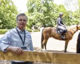 Thies Kaspareit, ein Mann mit Brille, steht vor einem Holzzaun. Im Hintergrund reitet eine Person auf einem Pferd.