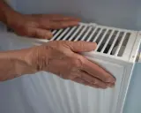 Manos sobre un radiador blanco, sintiendo el calor. El radiador hace ruido, quizás por el agua circulando.