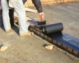 Pracownik nakłada masę bitumiczną na ceglaną ścianę, przygotowując ją do położenia hydroizolacji z rolki.