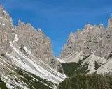 Vette rocciose imponenti sotto un cielo azzurro. Un sentiero scende verso il rifugio Giaf, tra pini e chiazze di neve.