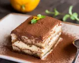 Pyszne tiramisu na biszkopcie, posypane kakao, z miętą i skórką pomarańczy. Obok łyżeczka i pomarańcza.
