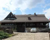 Drewniana bacówka Biała Woda z białym SUV-em na kamienistym podjeździe. Biało-czerwona flaga powiewa na maszcie.