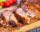 Une succulente épaule d'agneau rôtie, effilochée et servie sur une planche en bois, avec des tomates et de la laitue en arrière-plan.