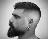 Mężczyzna z brodą i kolczykiem w nosie, z modnym, dłuższym buzz cut i cieniowanymi bokami.