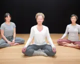 Tre kvinnor sitter i lotusställning på ett trägolv. De deltar i en klass i traumaanpassad yoga.