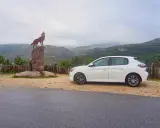 Ein weißer Kleinwagen parkt neben einer Wolfsstatue auf einem Felsen mit Blick auf eine bergige Landschaft. Tolle Carjet Erfahrungen für diese Reise!
