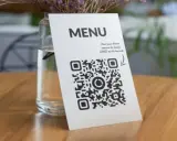 Un código QR de restaurante en una mesa de madera, junto a un jarrón con flores secas. Escanea para ver el menú.