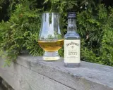 En liten flaska Jack Daniels Honey och ett whiskyglas står på en träbänk, omgivna av grönska.