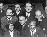 Un gruppo di uomini sorridenti, alcuni con baffi e occhiali, in una fotografia che evoca lo spirito irriverente del dadaismo.