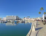 Boote im Hafen von Faro, Portugal, spiegeln sich im ruhigen Wasser. Palmen säumen die Promenade, wo Menschen in Cafés sitzen.