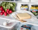 Ein Stück Käse liegt auf einem Holzbrett, daneben Radieschen und Gläser mit eingelegtem Gemüse. Verschiedene Käse sorten im Kühlschrank.