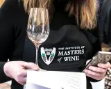 Eine Person mit einer Schürze des "Institute of Masters of Wine" hält ein Weinglas und ein Smartphone.