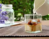 Milch wird in ein Glas mit Eiswürfeln und Kaffee gegossen, um einen erfrischenden Lavendel Latte zuzubereiten.