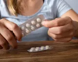 Dłonie trzymają opakowanie z tabletkami, które mogą być lekami z serotoniną. Na drewnianym blacie leży drugie, częściowo zużyte opakowanie.