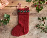 Röd stickad julstrumpa med grön misteldekoration och polkagrisar. En present och julgranspynt ligger på träbordet.