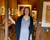 A woman smiles, opening the door to a photography west gallery filled with framed prints and display cases of artwork.