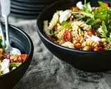 Zwei Schalen mit Nudelsalat mit getrockneten Tomaten, Pinienkernen und Rucola. Ein Löffel liegt in der vorderen Schale.