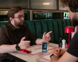 Robert Gryn, w okularach, gestykuluje podczas rozmowy przy stoliku w restauracji. Na stole stoi małe urządzenie z ekranem.