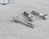 Trzy metalowe kotwy leżą na betonowej powierzchni. Każda kotwa ma gwintowany pręt i nakrętkę.