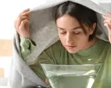 Kobieta z zasłoniętą głową kocem wykonuje inhalację na zatoki nad miską z gorącą wodą.