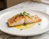 Ein saftiges Stück Backfisch im Airfryer, mit Kräutern und Gewürzen verfeinert, liegt auf einem Teller in flüssigem Gold.