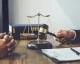 Dłoń trzymająca młotek sędziowski, waga sprawiedliwości i akta na drewnianym stole.
