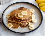 Placuszki bananowe na talerzu, ozdobione plasterkami banana. Idealne, gdy zastanawiasz się, co zrobić z bananów fit. Obok miseczka z dżemem.