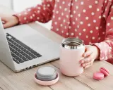 Kobieta przy laptopie trzyma kubek termiczny, obok leży nakrętka. Dowiedz się, jak wyczyścić nakrętkę od kubka termicznego, by napoje dłużej smakowały świeżo.