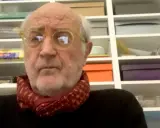 Ritratto di Francesco Pecoraro con occhiali gialli e sciarpa rossa, davanti a una libreria piena di scatole.