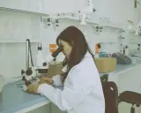 Kobieta w białym fartuchu pracuje przy mikroskopie w laboratorium. Na stole leżą próbki i sprzęt.