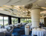 Salle élégante d'un restaurant chef de baie, tables dressées, vue sur la mer et jardin luxuriant. Plafond ondulé et luminaires modernes.