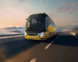 Autobús Setra amarillo brillante viaja por una carretera escénica al amanecer, con nubes y montañas de fondo.