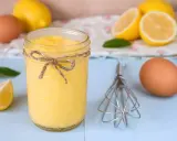 Un tarro de lemon curd casero, atado con cuerda, junto a limones, huevos y un batidor.