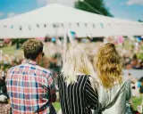 Tre vänner njuter av en solig dag på en musikfestival i Stockholm. En stor vit tältduk och flaggor syns i bakgrunden.