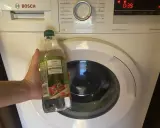 Ręka trzyma butelkę octu spirytusowego obok pralki Bosch. To początek domowego sposobu na mycie pralki octem.
