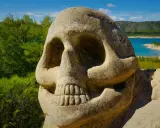 Escultura de calavera en la **ruta de las caras**, con un lago azul y colinas verdes al fondo.