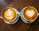 Zwei Cappuccinos mit Latte Art auf einem Holztisch. Sie bestehen aus Espresso, heißer Milch und Milchschaum.