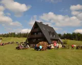 Drewniana bacówka z ludźmi siedzącymi przy stołach na trawie, w tle las i niebo z chmurami.