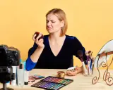 Donna con espressione disgustata annusa una crema viso chiusa, forse per verificarne la scadenza. Sul tavolo, trucchi e una telecamera.