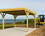 Un carport en bois est en cours de construction près de la mer. Une mini-pelle jaune est garée à côté, prête pour la suite des travaux. Une déclaration de carport est nécessaire pour ce projet.
