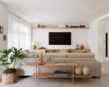 Estantería de madera con objetos decorativos, jarrones y cuadros. Un sofá beige y una planta grande complementan el espacio. Ideas de qué poner detrás del sofá.