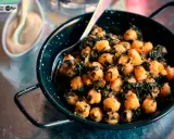Un plato de garbanzos con espinacas, servido en una sartén negra con un tenedor. Al fondo, una pequeña taza con una salsa cremosa.