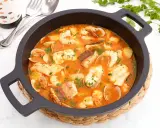 Una deliciosa cazuela de pescado con gambas, almejas y trozos de pescado blanco en una rica salsa roja, adornada con perejil fresco.