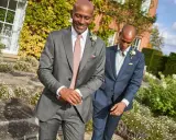 Two men in suits walk outdoors. The question "do you have to wear a suit to a wedding?" is answered by their stylish attire.