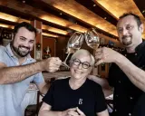 Dos hombres y una mujer brindan con copas de vino en Ca la Pepa, un restaurante acogedor con estanterías de vino.