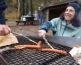 Vänner grillar korv vid en eldstad på Otto Pers gård. En kvinna ler glatt medan hon grillar.