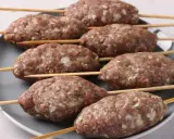 Siedem surowych kofta kebab na patyczkach, ułożonych na szarym talerzu, gotowych do grillowania.