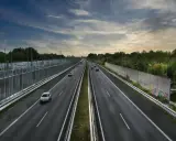 Szeroka autostrada z samochodami, wjazd na autostradę w tle, po bokach ekrany akustyczne i drzewa.